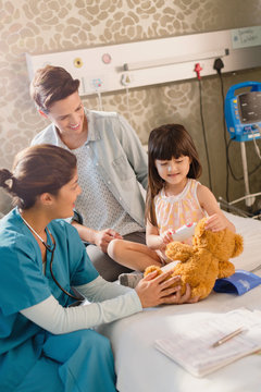 Female nurse and girl patient using digital thermometer on teddy bear in hospital room