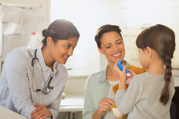 Female pediatrician and mother watching girl patient using inhaler in examination room