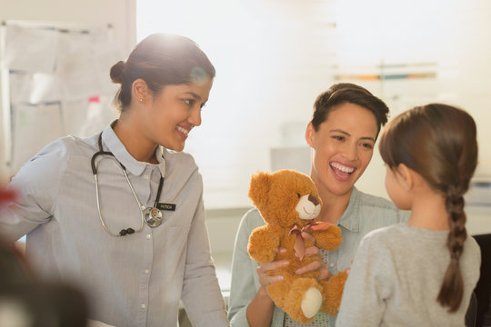Smiling female pediatrician and mother showing teddy bear to girl patient in examination room