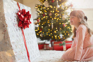 Girl smiling in anticipation at large Christmas gift near Christmas tree