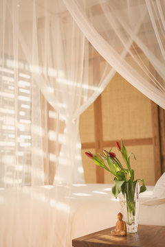 Tulip bouquet and Buddha statuette on bedside table next to canopy bed with white gauze curtains in tranquil bedroom