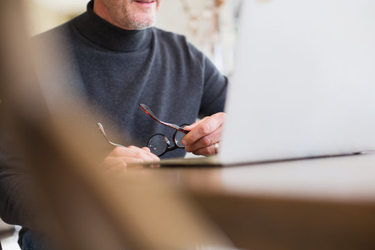 Mature male freelancer holding eyeglasses, working at laptop