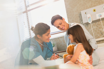 Smiling female nurse using stethoscope on girl patient in hospital room