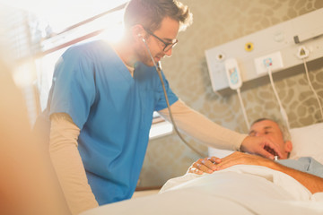 Male nurse using stethoscope on patient in hospital bed