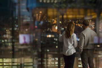 Businessman and businesswoman looking out urban office window at night