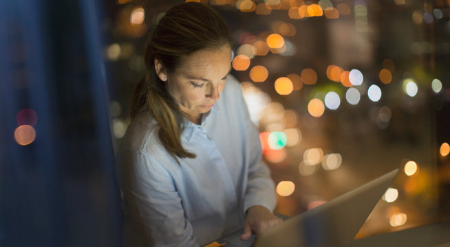 Serious, focused businesswoman working late at laptop in office window at night