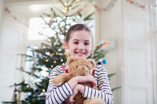 Portrait smiling, cute girl holding teddy bear in front of Christmas tree