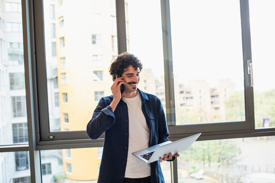 Man talking on smart phone and using laptop at urban apartment window