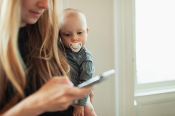 Curious baby girl with pacifier watching mother using smart phone