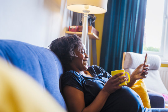 Happy Pregnant Woman Relaxing On Sofa With Tea And Smart Phone