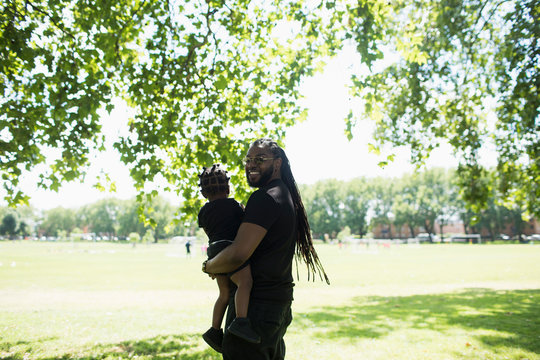 Portrait happy father with long braids carrying son in sunny park