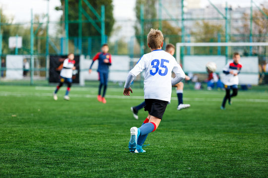 Boys in white and blue sportswear plays football on field, dribbles ball. Young soccer players with ball on green grass. Training, football, active lifestyle for kids concept
