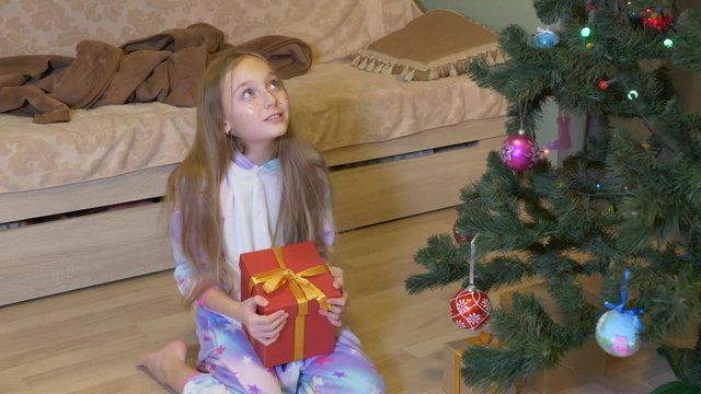 Adorable little girl opening christmas present at home. Disappointed girl in pajamas sitting near christmas tree and opening gift box with socks
