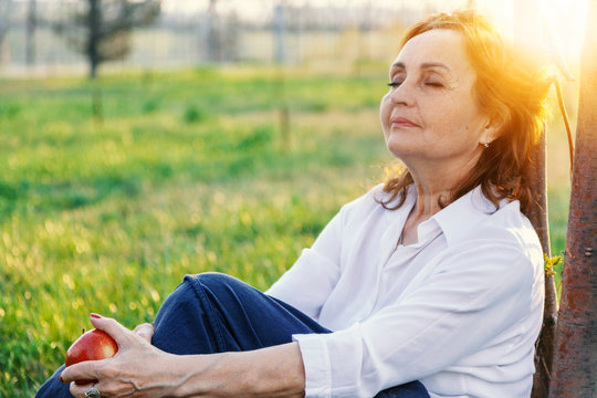 Happy senior woman relaxing on green lawn. Close up face of a mature brunette woman smiling relaxing. Retired woman in casuals sitting outdoor in a summer day.