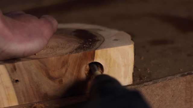 Close up of a carpenter drilling a circular hole into a large slab of hardwood, held down by the carpenter's bare hand