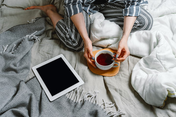 Faceless portait of woman sitting in her bed with tablet and tea, selective focus