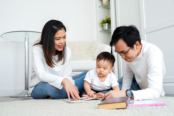 Happy modern Asian parents sitting on floor with cute infant boy and reading fairy tale while enjoying time together at home