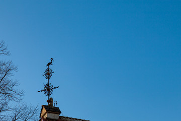 Background image - a metal weather vane in the shape of a stork sitting on a base supported by a stone lion against a clear blue sky