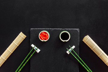 Table served for eating sushi. Chopsticks, small bowls with ginger and sause, mat on black background top-down copy space