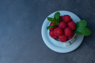  Raspberries with mint leaves in a white mug. Two little pieces of sugar. Dark gray stone table. View from above. Copy space, place for text.