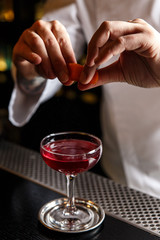 Bartender prepairing a cocktail at the bar, squeezing an orange peel over a drink in a cocktail glass