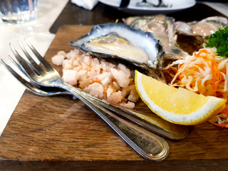 Two stainless steel fork with lime sliced, vegetable, salt and new Zealand oysters blurred background on wooden tray at restaurant. Fresh seafood and Selected focus at fork.
