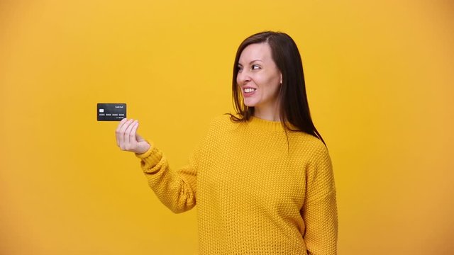Smiling beautiful young woman in bright sweater shirt posing isolated over yellow orange background in studio. People lifestyle concept. Hold in hand credit bank card, take give in hand and receive.