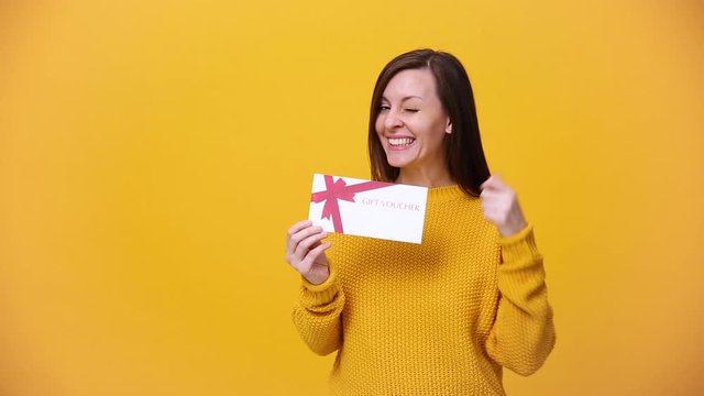 Smiling beautiful young woman in bright sweater posing isolated over yellow orange background in studio. People sincere emotions, lifestyle concept. Hold in hand gift certificate, card coupon voucher.
