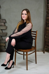 Fototapeta na wymiar Full-length photo of a pretty brunette girl with excellent make-up in dark clothes sitting on a stand on a gray background in a luxury vintage interior. The concept of a glamorous portrait.
