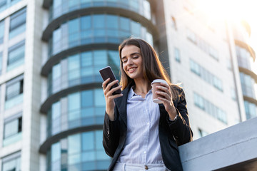 Business woman with coffe and talking on the phone near office. Young woman with smartphone standing against street blurred building background. Beautiful girl in suite with phone and cup of coffee