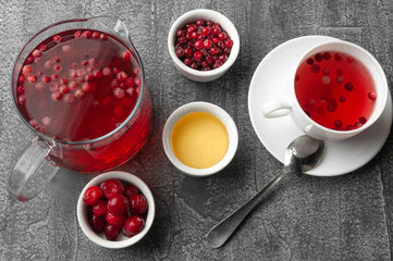 Glass jug with a drink of cranberries and cherries. Next to the jug are plates with berries, honey and a white porcelain cup. View from above. On a gray concrete background.