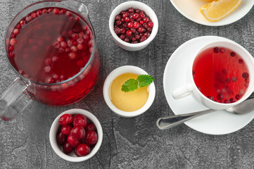 Glass jug with a drink of cranberries and cherries. Next to the jug are plates with berries, honey and a white porcelain cup. View from above. On a gray concrete background.