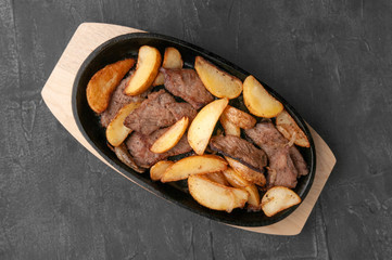 Fried potato slices with onions and beef. In a cast-iron pan with a wooden stand. View from above. Gray concrete background.