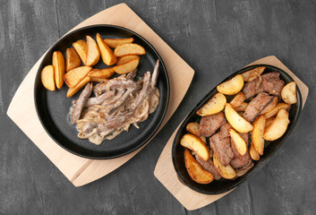 Fried potato slices with onions, beef and beef stroganoff. In a cast-iron pan with a wooden stand. View from above. Gray concrete background.