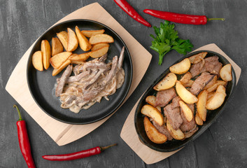 Fried potato slices with onions, beef and beef stroganoff. In a cast-iron pan with a wooden stand. Decorated with chili peppers and parsley. View from above. Gray concrete background.