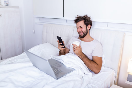 Young Businessman Working Happy And Relaxed From Bed With Laptop Computer Using Mobile Phone In Workaholic Concept , Working From Home In The Morrning And Drinking Coffe