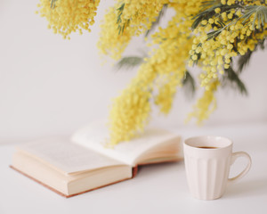 Cup of coffee and a book on white desk with a bouquet of mimosa.  feminine flat lay. Cozy breakfast. Easter, spring concept
