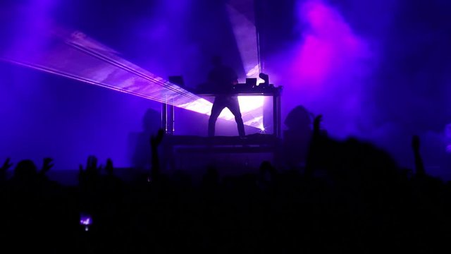 Flashing purple pink lights on stage of DJ performing pop music at a youth festival in front of large crowd attending the night show
