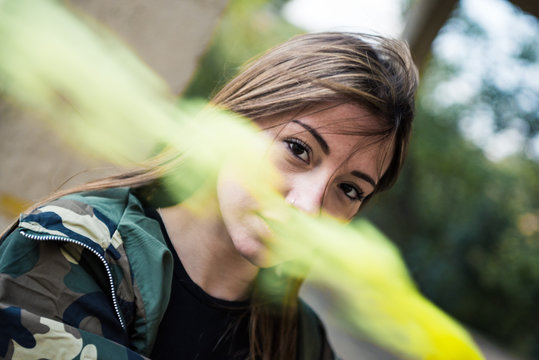 Brown hair girl holds a smoke bomb that throws yellow smoke around. Young girl wears an urban style, military jacket and piercing.