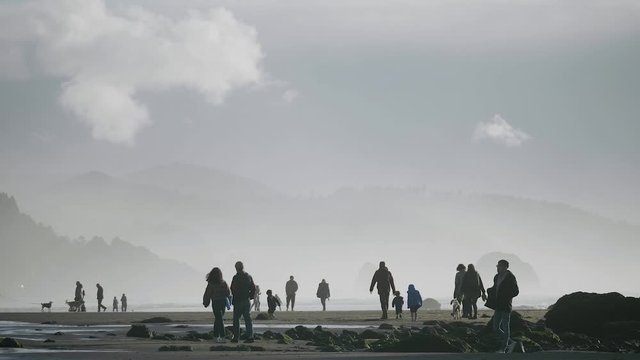 Many people silhouette walking on sandy beach at Cannon Beach Oregon on a moody day with mountains in background slow motion 