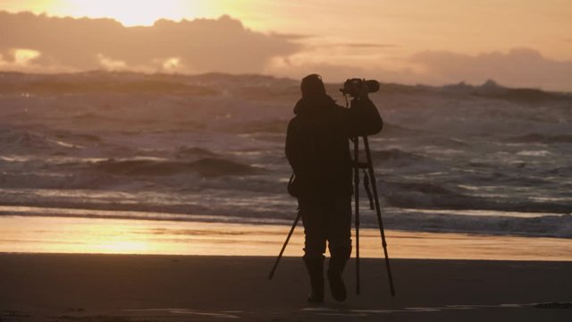 Male photographer silhouette sunset sky beach ocean crashing waves leaving slow motion holding camera and tripod
