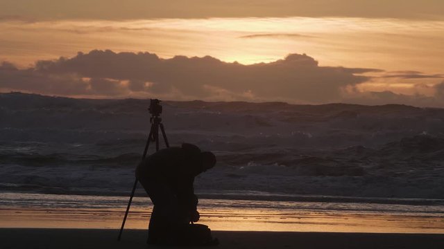 Male photographer silhouette sunset sky sandy beach ocean crashing waves checking bag changing lenses slow motion