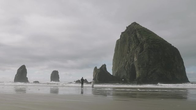 Couple people standing on sandy pacific ocean beach in front of huge haystack rock cliff at cannon beach oregon 