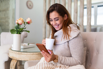 Pretty young woman sitting on sofa and enjoying first morning coffee in cozy apartment. Smiling woman sitting on sofa while drinking hot tea. Happy girl relaxing at home on a bright winter morning.