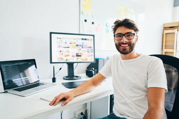 Young photographer looking at camera and smiling while sitting in office with laptop editing pictures