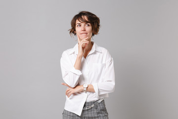 Pensive young business woman in white shirt posing isolated on grey wall background in studio. Achievement career wealth business concept. Mock up copy space. Put hand prop up on chin, looking aside.