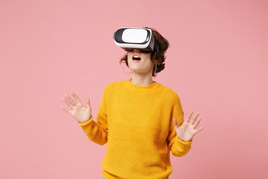 Amazed young brunette woman girl in yellow sweater posing isolated on pink wall background. People lifestyle concept. Mock up copy space. Watching in headset of vr virtual reality, spreading hands.