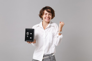 Joyful young business woman in white shirt isolated on grey background. Achievement career wealth business concept. Mock up copy space Doing winner gesture hold metal bank safe for money accumulation.