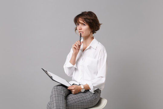 Pensive young business woman in white shirt isolated on grey background. Achievement career wealth business concept. Mock up copy space. Hold clipboard with papers document, writing notes, sitting.