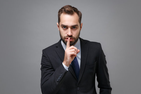 Young business man in classic suit shirt tie posing isolated on grey background. Achievement career wealth business concept. Mock up copy space. Saying hush be quiet with finger on lips shhh gesture.